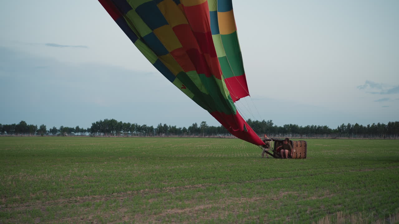 passengers crouching beside deflating colorful hot air balloon envelope dragging heavy fabric down to ground across expansive green field under pale sky