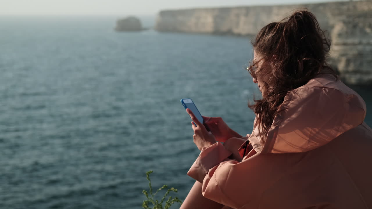 mujer usando el teléfono en un acantilado costero con vistas al océano