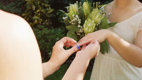 Happy-diverse-female-couple-holding-bunch-of-flowers-and-wearing-ring-in-garden