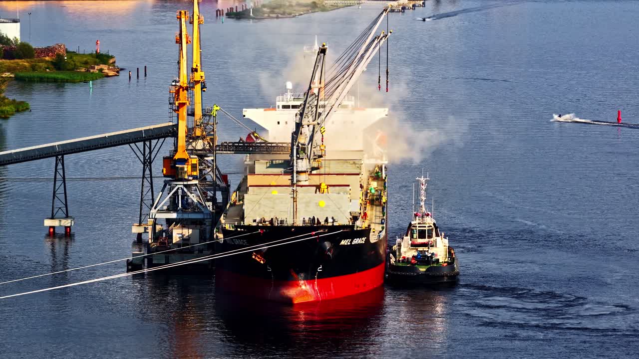 Cargo ship being loaded at an industrial port with cranes and a tugboat assisting alongside, with smoke rising from the vessel and calm blue water surrounding the scene