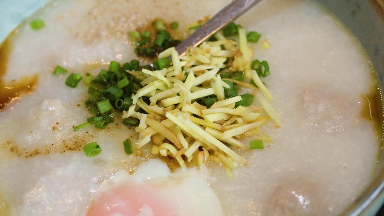 Close-up bubur sarapan tradisional Asia dengan taburan jahe, daun bawang, dan telur dalam mangkuk.