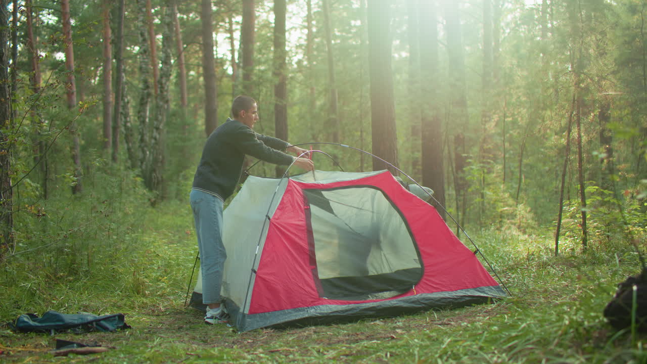 young man carefully dismantles red and gray camping tent in forest clearing while woman assists from inside as sunlight filters through trees and backpack rests on ground near tent