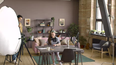 Woman-Sitting-On-A-Table-With-Laptop-And-Microphone-To-Recording-A-Podcast-While-The-Man-Is-Adjusting-The-Camera