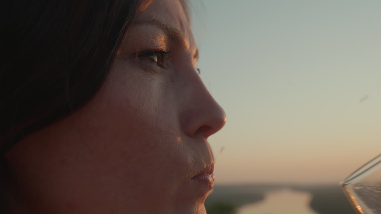 Side profile of woman sipping red wine from glass as tiny insects hover around in warm sunset glow, blurred river landscape and golden light reflecting in liquid creating serene outdoor moment