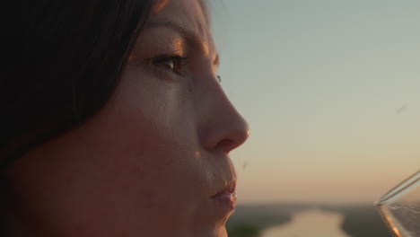side profile of woman sipping red wine from glass as tiny insects hover around in warm sunset glow, blurred river landscape and golden light reflecting in liquid creating serene outdoor moment