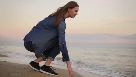 Attraktive-Junge-Frau-Sitzt-Am-Meer-Und-Versucht-Bei-Sonnenuntergang-Mit-Der-Hand-Am-Strand-Die-Wassertemperatur.-Zeitlupe