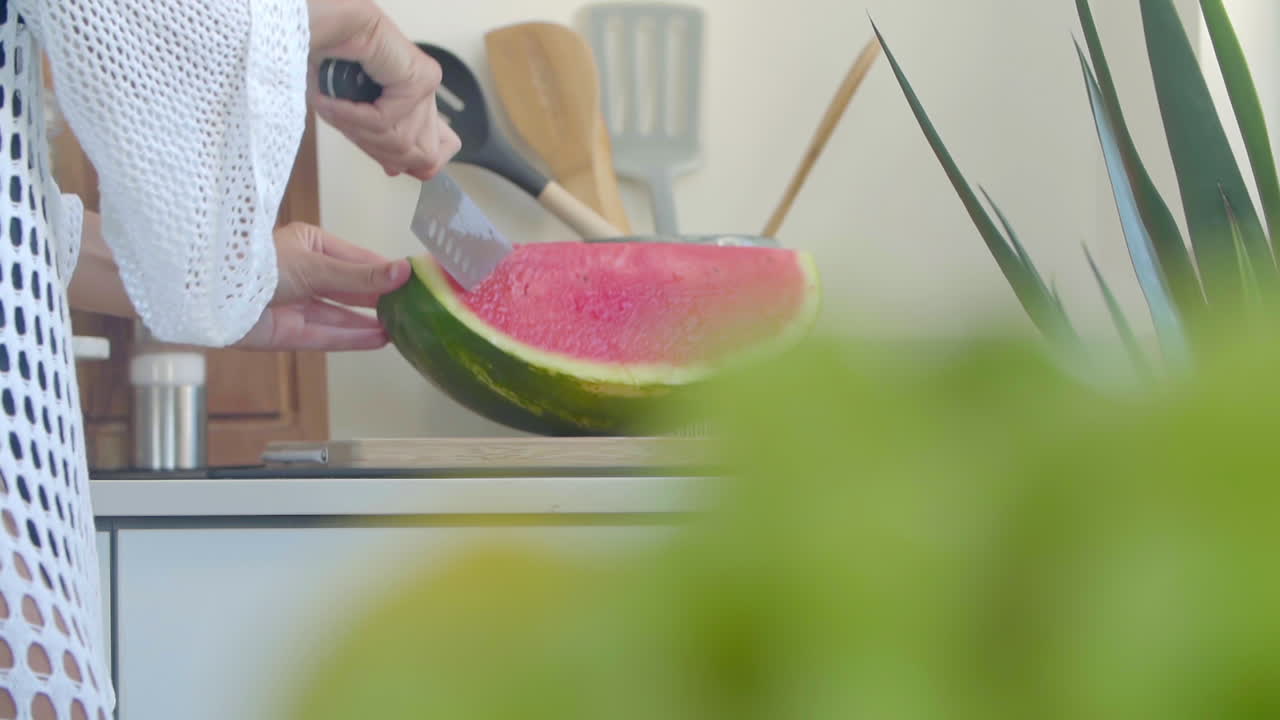 cerca de una mujer en un vestido blanco corta una sandía madura en una tabla de madera con un cuchillo grande en la cocina