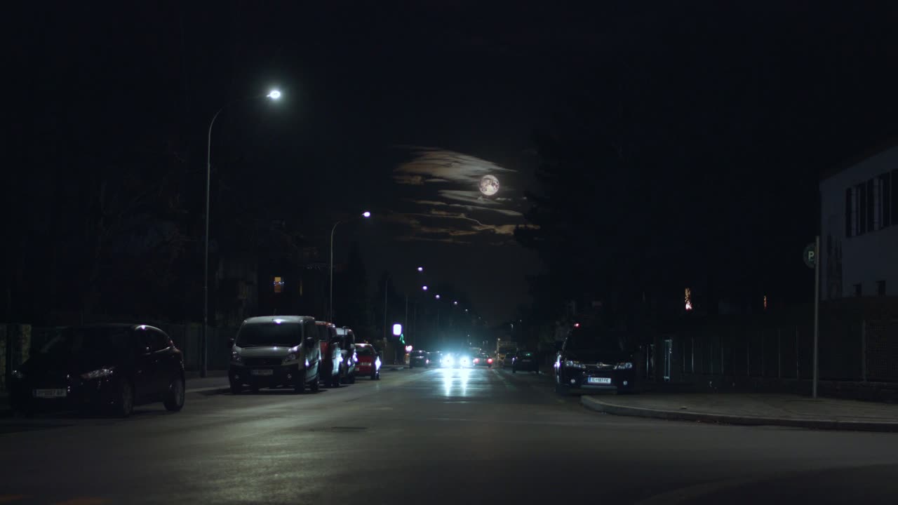Cars driving through a dark street at night with full moon and clouds in the background