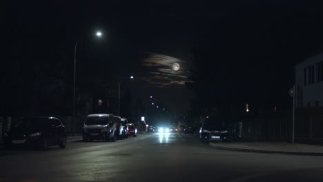 cars driving through a dark street at night with full moon and clouds in the background
