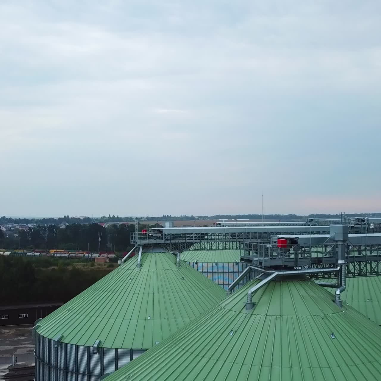 Rising along a huge silver metal granary tank standing at the territory of modern silo plant. Look at the slope roofs of elevators at the backdrop of farmlands