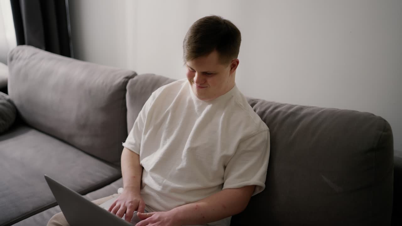 Man with down syndrome at home sits on sofa, typing on laptop