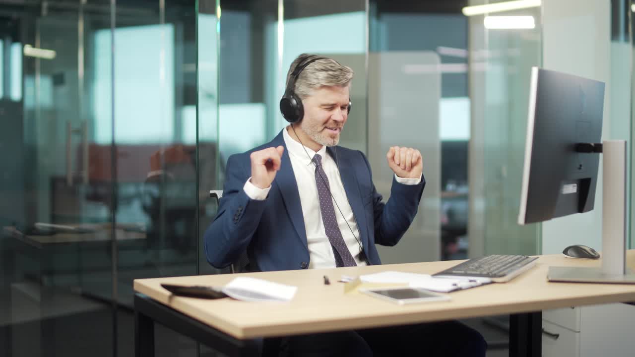 Uomo d'affari maturo che ascolta musica in ufficio alla scrivania del computer. Un anziano uomo d'affari allegro con i capelli grigi, con le cuffie, si rilassa, balla e canta.