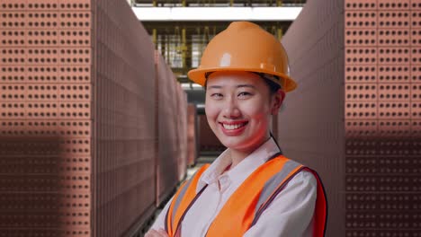close up side view of asian female engineer with safety helmet crossing her arms and smiling to camera while standing with red brick packed in stacks are stored