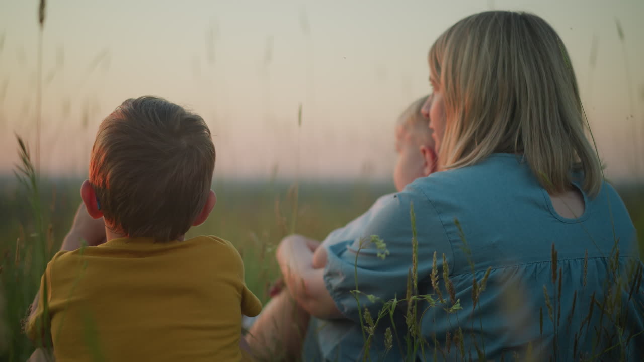 해가 지는 동안 들판에 앉아있는 파란 옷을 입은 어머니의 클로즈업 백, 어린 아이를 무에 들고 부드럽게 움직이고, 큰 아이는 모두 호수를 바라보면서 에 앉습니다.