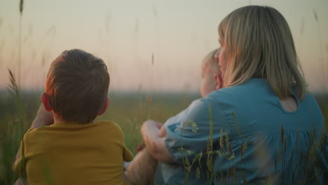 close-up back shot of a mother in a blue gown sitting in a field during sunset,gently moving while holding her younger child on her lap.the older child sits beside her as they all gaze out at the lake