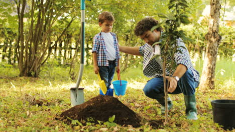 Portrait of a little boy and his dad planting a tree. Man takes the tree from the bucket and puts it into the hole. Then he shows and explains something to his son. Then they smile. Blurred background