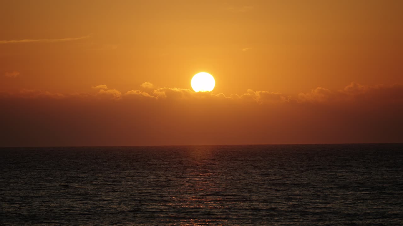 The sun sets above a blanket of clouds, casting intense golden hues over the calm Atlantic Ocean