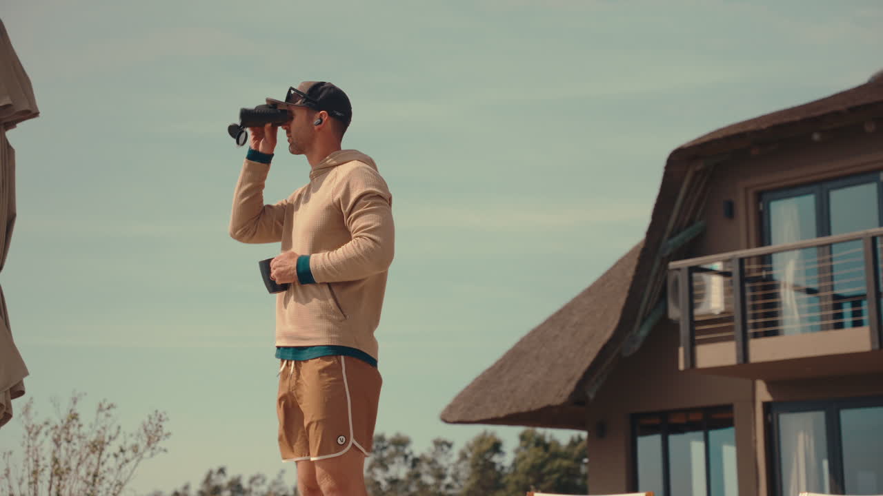 Man surveying landscape with binoculars, then sips coffee, outside of modern home on sunny windy day, static 4k birdwatching on African Safari