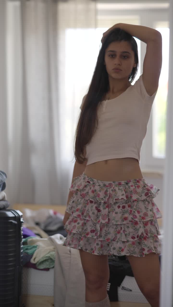 Young woman getting ready in front of a mirror with clothes in a messy bedroom