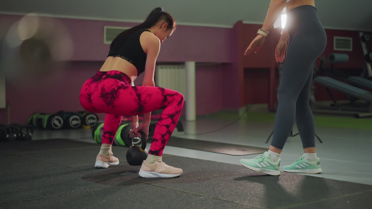 Dos mujeres practicando peso muerto con kettlebell juntas. El entrenador corrige la postura y el agarre. Suelo de colchonetas de gimnasio, luz de estudio suave, asistencia de compañero. Un ambiente de entrenamiento instructivo y colaborativo, enfocado en...