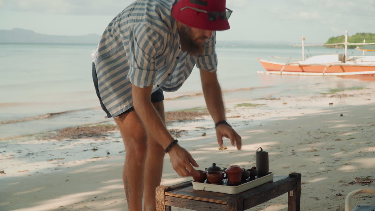 Allestimento del tè in spiaggia con un uomo che sistema un set portatile su un tavolino basso, cappello rosso, barca sullo sfondo, marea sulla riva, pantaloncini estivi casual, stile di vita spontaneo, mani che posizionano teiera e tazze.