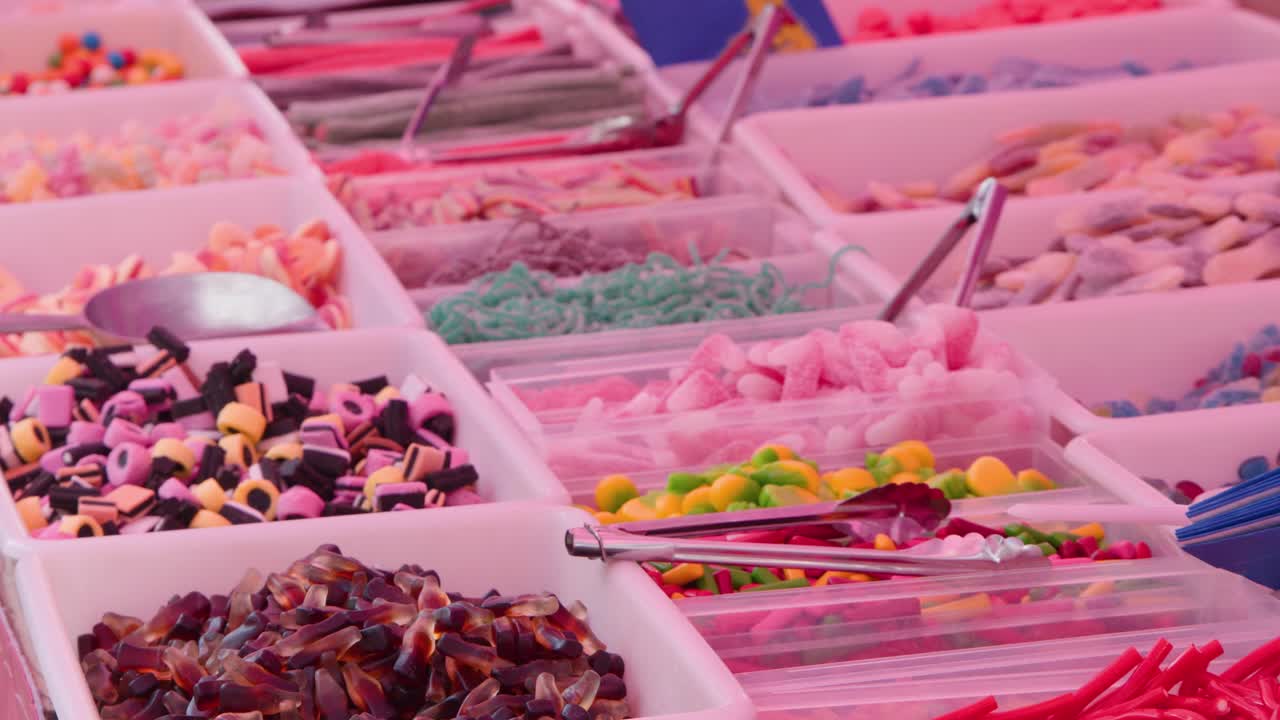 Surtido de caramelos expuestos en recipientes en un mercado al aire libre lleno de vida, con la cámara desplazándose lentamente por los dulces