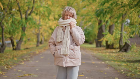 mature woman wearing soft pink jacket and thick white scarf adjusts muffler with one hand while other hand stays in pocket, walking along peaceful autumn path surrounded by golden foliage