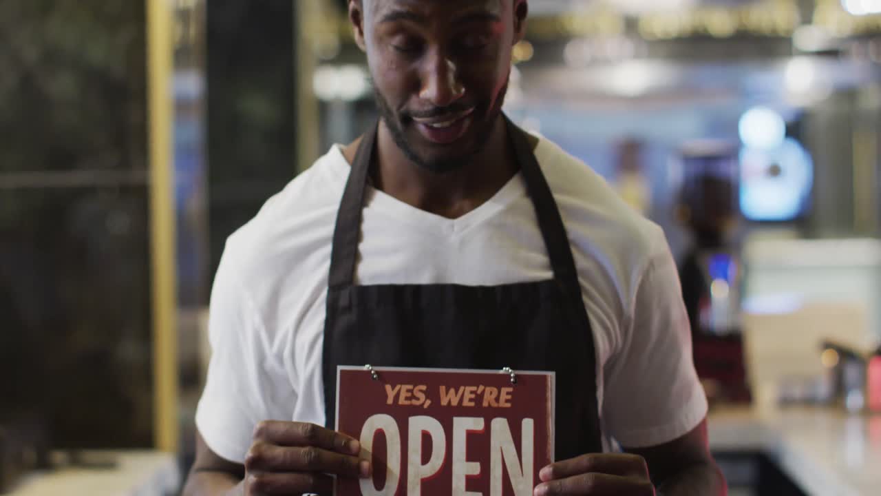 Portrait of happy african american barista holding 'we're open' sign in cafe
