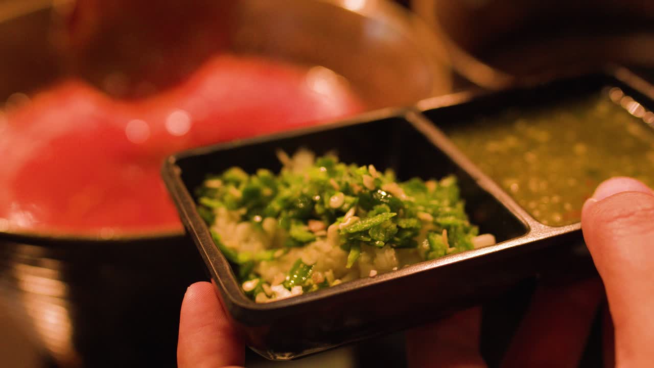 Hand presents divided condiment tray above simmering hotpot broth, warm lighting, close-up perspective