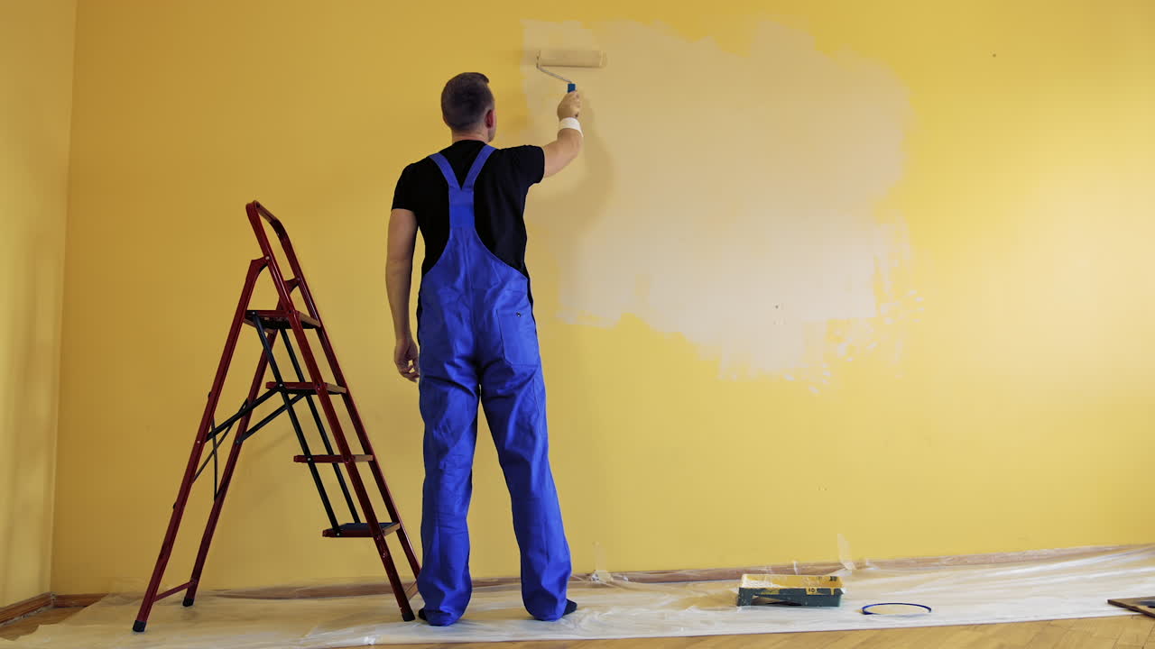 Room decoration with new color. Professional worker in blue overalls painting wall with a paint roller in empty room. Rear view.