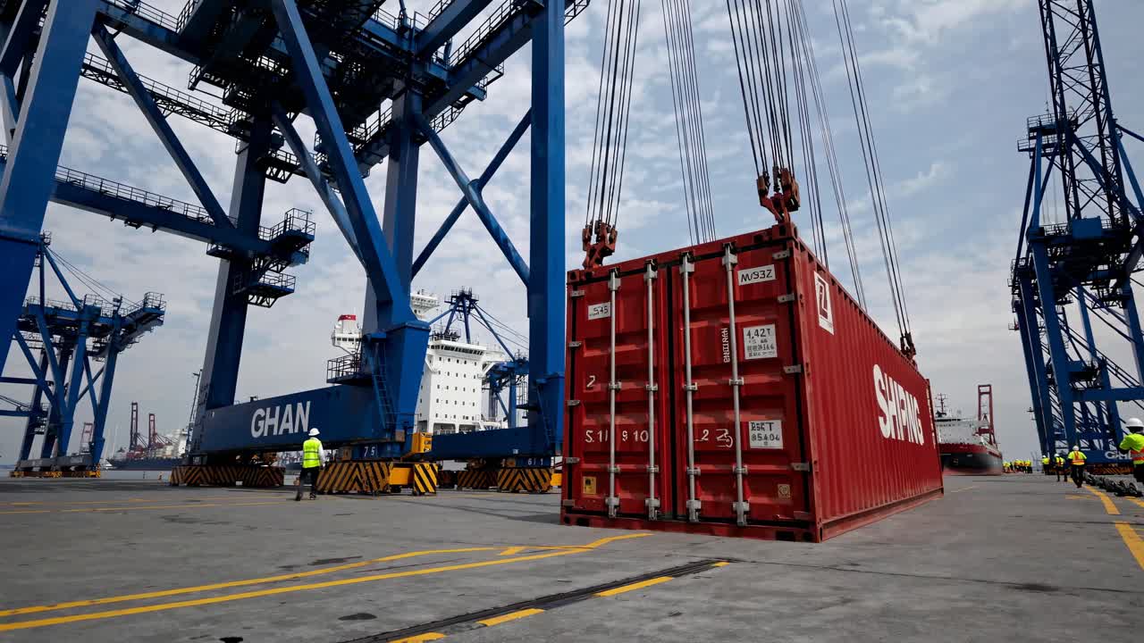 Low-angle video shot of a red shipping container being lifted by a crane at a busy port