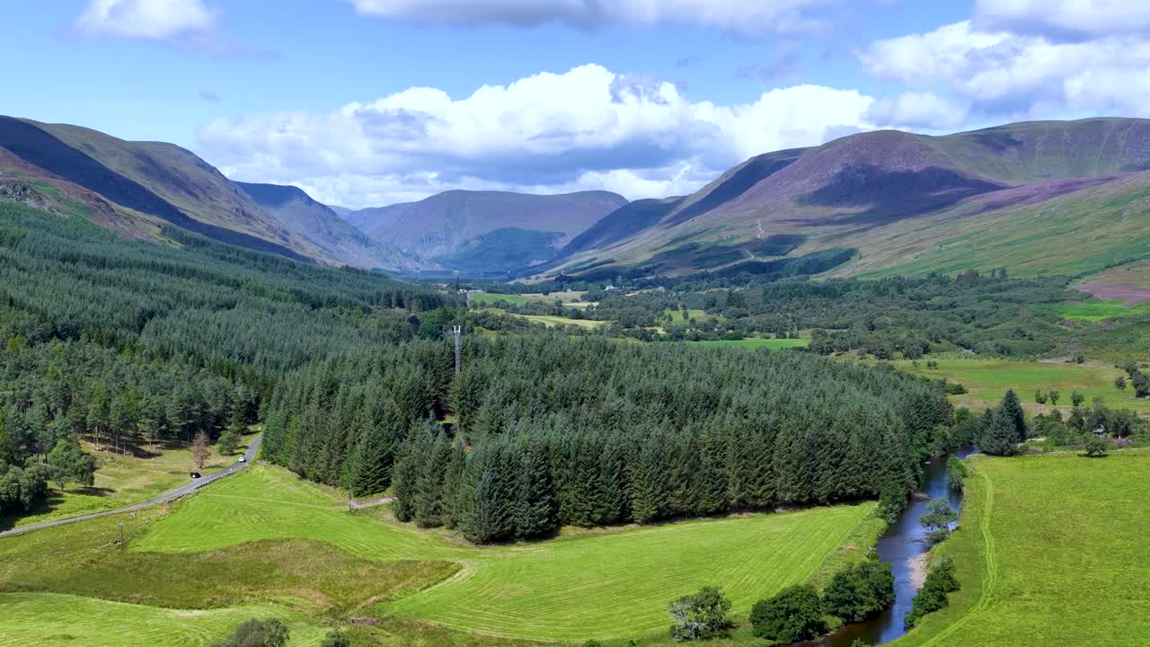 Drone glides above green river valley, dense forest, and mountains under bright daylight skies