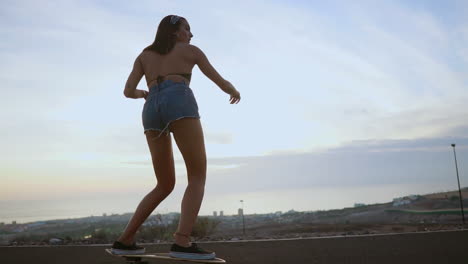 In-slow-motion,-a-stylish-young-skateboarder-gracefully-rides-her-board-in-shorts-at-sunset-along-a-mountain-road,-offering-a-breathtaking-view-of-the-mountains