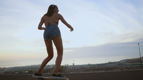 en cámara lenta, una elegante joven patinadora graciosamente monta su tabla en pantalones cortos al atardecer a lo largo de un camino de montaña, ofreciendo una vista impresionante de las montañas