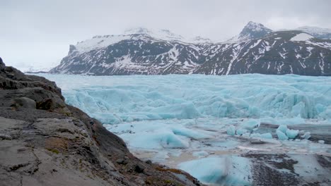 static, shot over the rocky coast, of the turquoise, skaftafellsjokull glacier, snowy mountains in the background, on a cloudy day, in south coast of iceland