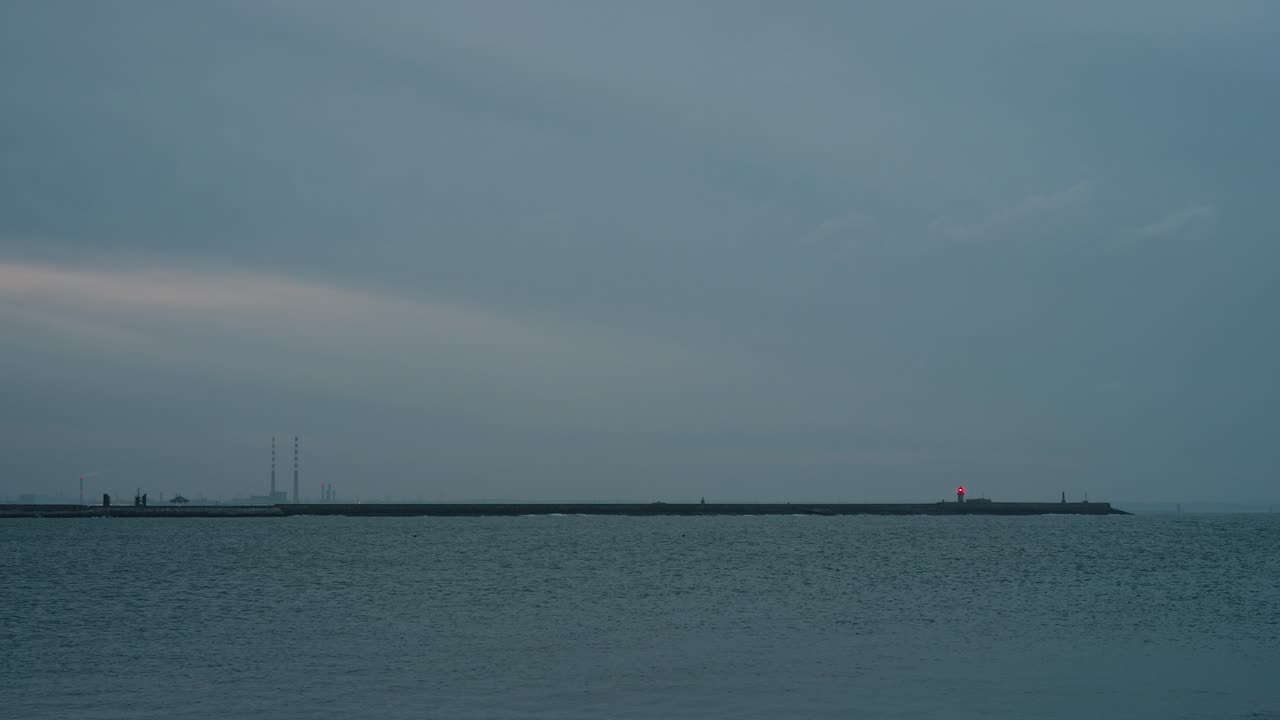 Dublin Bay at dusk with Poolbeg Towers and East Pier Lighthouse visible in the distance. The calm sea reflects the soft blue hues of the sky.
