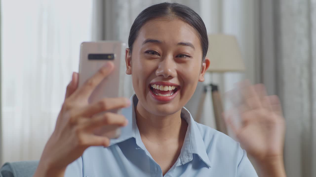 close up de una mujer asiática agitando la mano haciendo una llamada de video en un teléfono inteligente mientras está sentada en el sofá en la sala de estar