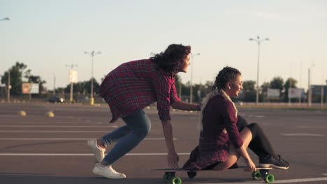 Vista-Trasera-De-Una-Mujer-Sentada-En-Un-Longboard-Mientras-Su-Amiga-La-Empuja-Hacia-Atrás-Y-Corre-Durante-La-Puesta-De-Sol