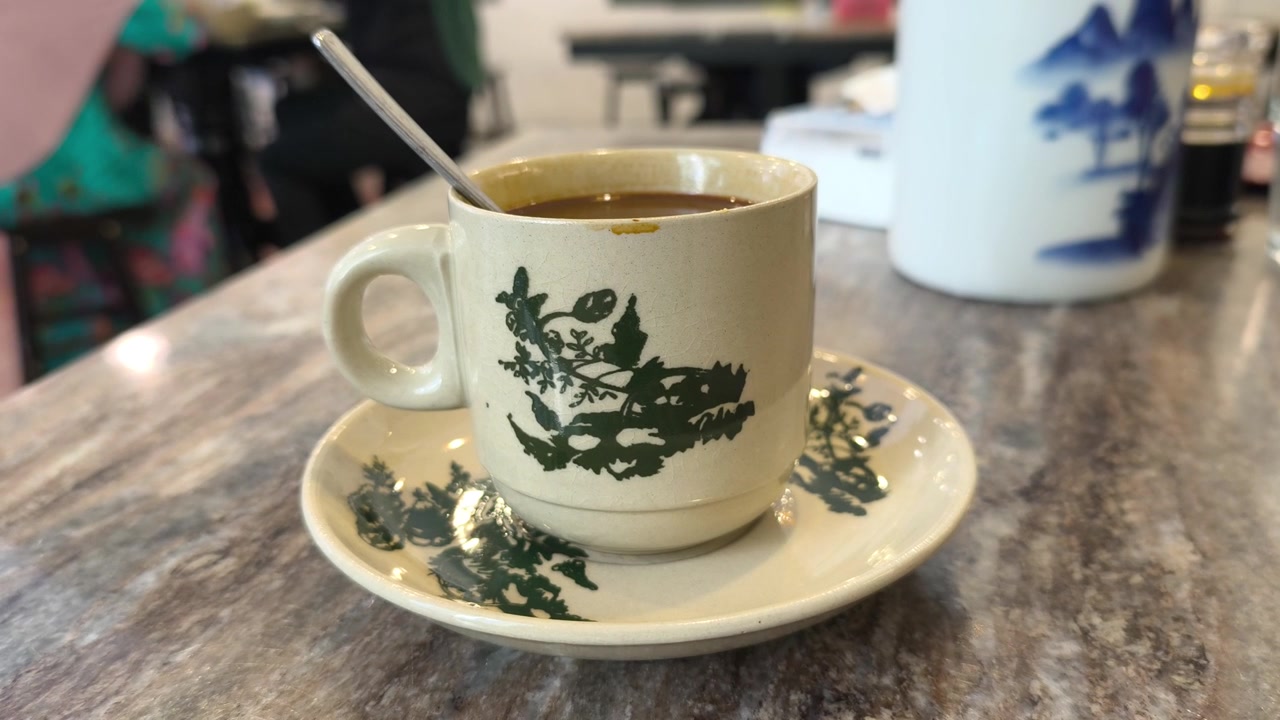 Una tazza di caffè al latte cremoso è servita in una tradizionale tazza e piattino di ceramica. Questa preparazione classica riflette l'autentica cultura kopitiam della Malesia. Emana vapore delicatamente su un tavolo rustico.