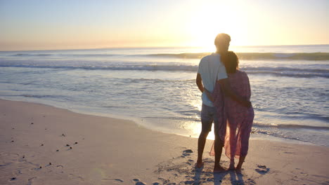 Biracial-Paar-Umarmt-Sich,-Während-Es-Einen-Sonnenuntergang-Am-Strand-Mit-Kopierraum-Beobachtet