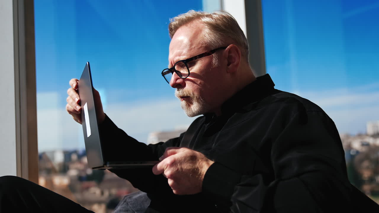 Concentrated male in black outfit sits ay window holding a laptop. Man in glasses checks e-mail and closes his gadget.