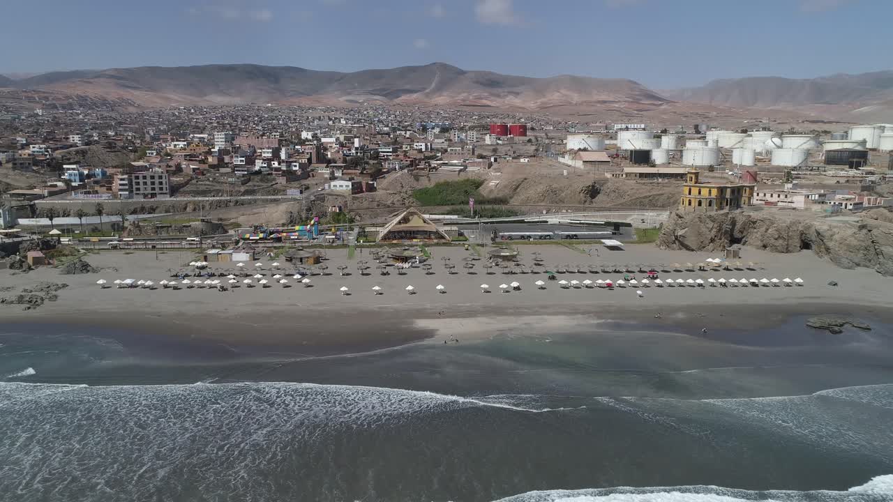 Dynamic aerial view of a coastal city with a sandy beach, lined umbrellas, and industrial structures. Perfect for travel blogs, urban studies, and stock imagery. Mollendo, Arequipa, Peru.
