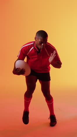 vertical video of african american male rugby player running with rugby ball over pink lighting
