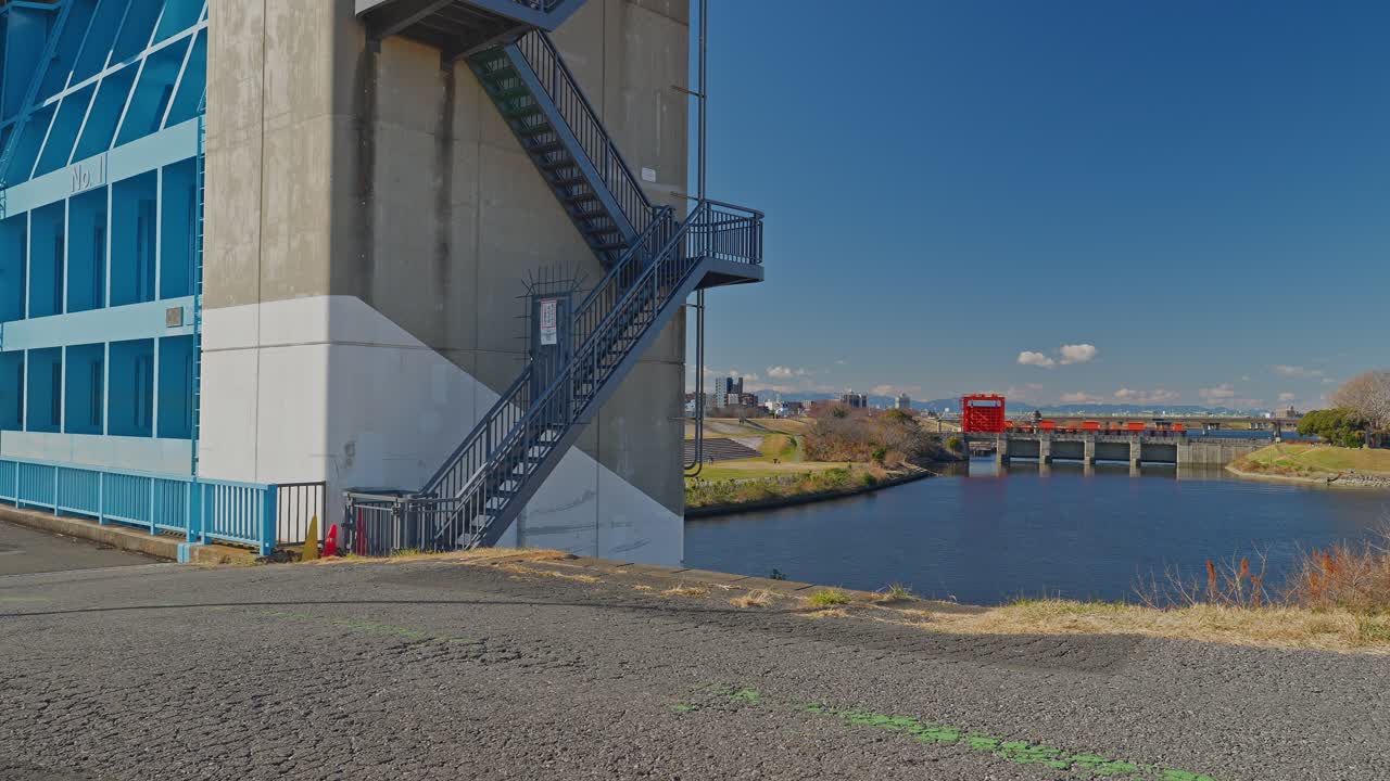 A detailed shot of the modern Blue Iwabuchi Sluice Gate's concrete tower and steel staircase. In the distance, the historic Red Sluice Gate is visible across the calm water of the Arakawa river system