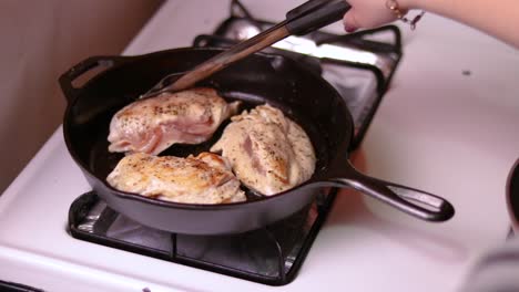 una vista en primera persona de tres piezas de pechuga de pollo que se fríen en una sartén forjada encima de una estufa de gas
