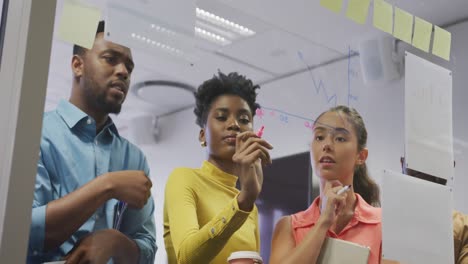 Diverse-male-and-female-business-colleagues-taking-notes-on-glass-wall-and-talking-in-office