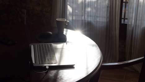 a shot of a laptop and a steaming cup of coffee on a circular wooden table near a window with curtains at sunrise