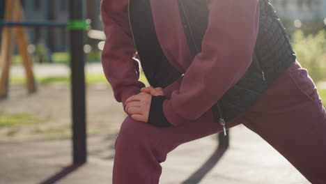 female figure in hoodie and quilted vest performs forward lunge with hands braced on leg under bright sun casting long shadows across modern park walkway with dew kissed blooming flowers