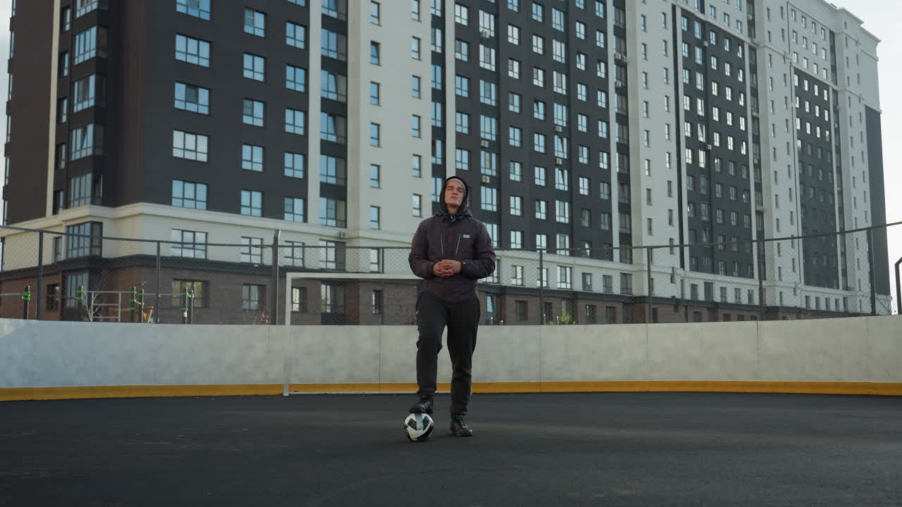 joven está en equilibrio con un pie en la pelota de fútbol, las manos juntas, mientras gira la cabeza y el brazo, respaldado por edificios urbanos de gran altura en una arena deportiva soleada
