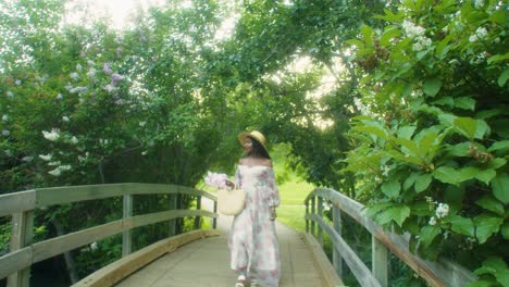 black woman with picnic basket walking over bridge smelling flowers lilacs wide pan close up